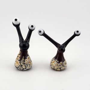 May include: Two glass snail figurines with black bodies and long stalks topped with white and black eyes. The base of each snail features a mottled pattern of cream, tan, and dark brown spots, creating a textured appearance on the smooth glass surface.