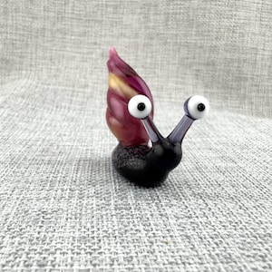 May include: A glass snail figurine featuring a dark black body with a speckled texture and two protruding stalks topped with white and black eyes. The snail shell is a swirling blend of deep purple, magenta, and yellow tones in a smooth finish.