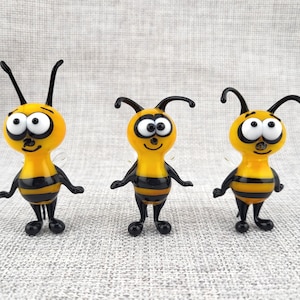Peut inclure: Trois figurines d'abeilles en verre alignées. Chaque abeille possède une tête et un corps jaunes avec des rayures horizontales noires, de grands yeux blancs à pupilles noires, des antennes noires, ainsi que des bras et des jambes noirs.