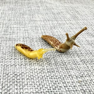 May include: Two glass slug figurines. One slug is bright yellow with brown spots on its back. The second slug is a translucent tan color with darker brown mottled patterns and two prominent, elongated eye stalks extending from its head.