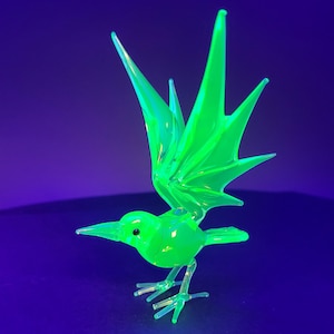May include: A vibrant neon green glass bird figurine with outstretched wings, a pointed beak, and thin legs. The translucent material glows intensely under ultraviolet light, highlighting the sharp, angular details of the wings and body structure.