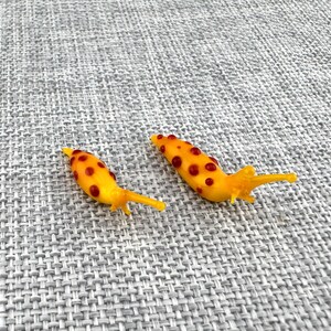 May include: Two small, yellow, slug-shaped figurines made of glass. Each figurine features a smooth, elongated body with a series of raised, dark red, circular spots distributed along the top and sides, and a pointed tail end.