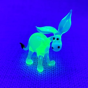 Puede incluir: Una pequeña figura de cristal con forma de burro que brilla en un tono verde neón intenso bajo luz ultravioleta. El burro tiene orejas grandes y erguidas, un cuerpo redondeado, cuatro patas y una cola pequeña con la punta oscura y ojos negros.