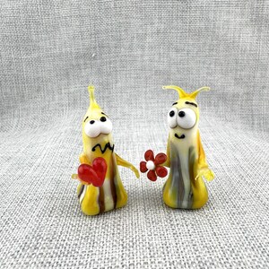 Peut inclure: Deux figurines en verre en forme de limaces stylisées avec des corps rayés jaunes et blancs. Une limace tient un petit cœur rouge, tandis que l'autre tient une fleur rouge avec un centre blanc. Elles ont de grands yeux blancs et noirs.
