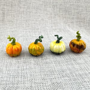 May include: Four small glass pumpkin figurines arranged in a row. The first is bright orange, the second is yellow-green with darker speckles, the third is pale yellow, and the fourth features a marbled brown and orange pattern. Each has a green stem.