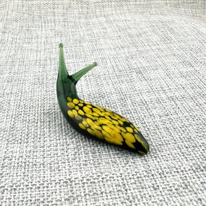 May include: A glass slug figurine featuring a translucent green head and two upright antennae, with a black body covered in a pattern of irregular, bright yellow spots that extend from the midsection down to the tapered tail end.