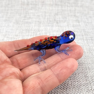 Op de afbeelding: Een klein glazen hagedisbeeldje met een diepblauw lichaam en oranje en bruine gevlekte patronen op de rug. Het wezen heeft een rij verhoogde rode stippen langs de ruggengraat, vier doorschijnende blauwe poten en één wit oog met een zwarte pupil.