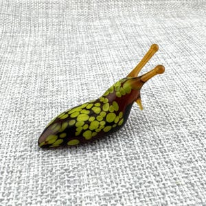 Könnte beinhalten: Eine Glasfigur in Form einer Nacktschnecke mit dunkelbrauner Grundfarbe und einem Muster aus hellen, lindgrünen Punkten. Die Schnecke hat zwei längliche, durchscheinende, bernsteinfarbene Fühler, die aus ihrem Kopf ragen, und eine glatte, glänzende Oberfläche.