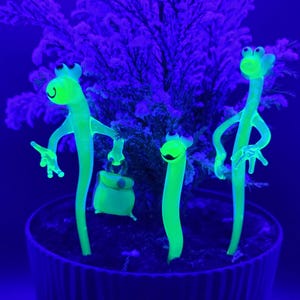 Puede incluir: Tres figuras de vidrio de color verde neón que brillan en la oscuridad, con forma de criaturas esbeltas de cuello largo, ojos grandes y extremidades delgadas. Una figura sostiene un bolso rectangular pequeño. Están clavadas en tierra oscura.