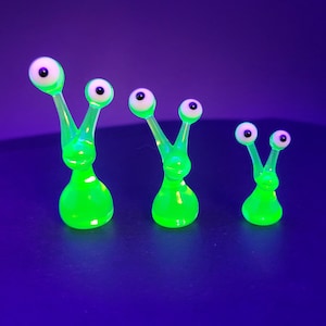 Può includere: Tre statuette in vetro verde neon brillante a forma di creature con due lunghi steli sormontati da occhi bianchi e neri. Le figure sono disposte in fila dalla più grande alla più piccola, ognuna con una base arrotondata e una superficie liscia e traslucida.