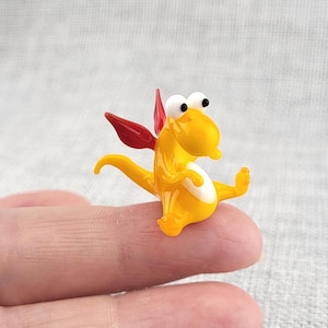 May include: A small glass figurine of a yellow dragon with a white belly, red wings, and large white eyes with black pupils. The creature has a curved tail and is depicted in a seated position with its arms and legs extended outward.