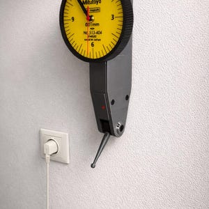 Puede incluir: Un reloj indicador de cuadrante Mitutoyo con esfera amarilla y bisel negro está montado en una pared blanca. El reloj muestra la hora y tiene un cuerpo negro y gris. El reloj está enchufado a una toma de corriente.