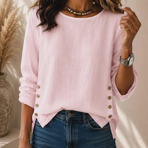May include: A light pink long-sleeve top featuring a round neckline and a row of four small brown buttons running vertically along each side of the hem. The garment is paired with dark blue denim jeans, creating a simple and clean look.