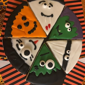 May include: A set of five triangular cookies arranged in a circle, decorated with icing to resemble Halloween characters including a pumpkin, vampire, monster, mummy, and ghost. The cookies are presented on a black and orange striped cardboard base.