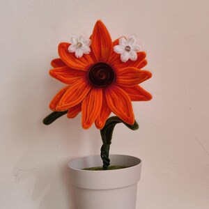 Può includere: Un fiore artigianale realizzato con scovolini arancioni e un centro marrone scuro, con due piccoli fiori in tessuto bianco applicati sui petali superiori. Il fiore ha uno stelo verde ed è piantato in un vaso di plastica grigio chiaro.