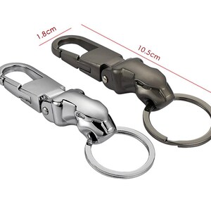 May include: Two metal keychains featuring a jaguar head design, one in a polished silver finish and one in a dark gunmetal gray finish. Each keychain includes a spring-loaded clasp and a circular ring, with dimensions of 1.8 cm width and 10.5 cm length.