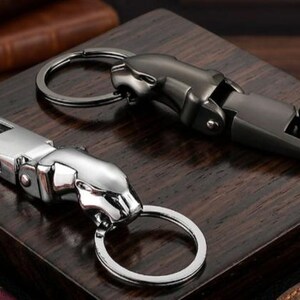 May include: Two metal keychains featuring a stylized jaguar head design. One keychain is finished in a polished silver tone, while the other is finished in a matte gunmetal gray. Each piece includes a spring-loaded clasp and a circular ring.