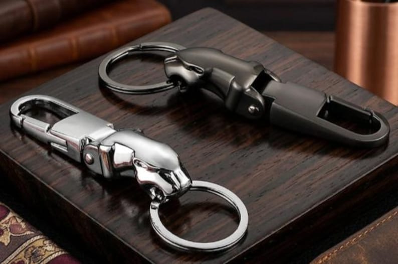 May include: Two metal keychains featuring a stylized jaguar head design. One keychain is finished in a polished silver tone, while the other is finished in a matte gunmetal gray. Each piece includes a spring-loaded clasp and a circular ring.