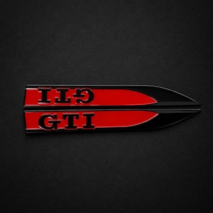 Puede incluir: Dos emblemas automotrices horizontales y alargados con una franja central roja brillante bordeada por bordes negros. Cada emblema muestra las letras GTI en una fuente negra, negrita y sin remates. El diseño termina en punta a la derecha.