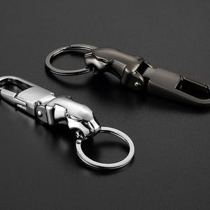 May include: Two metal keychains featuring a stylized jaguar head design. One keychain is finished in a polished silver tone, while the other is finished in a dark gunmetal gray. Each includes a spring-loaded lobster clasp and a circular ring.