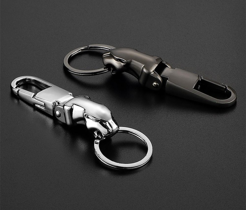 May include: Two metal keychains featuring a stylized jaguar head design. One keychain is finished in a polished silver tone, while the other is finished in a dark gunmetal gray. Each includes a spring-loaded lobster clasp and a circular ring.