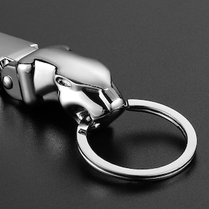 May include: A polished silver-tone metal keychain featuring a stylized jaguar head sculpture. The jaguar head is connected to a circular split ring for keys. The metallic surface has a reflective finish with smooth, contoured features.