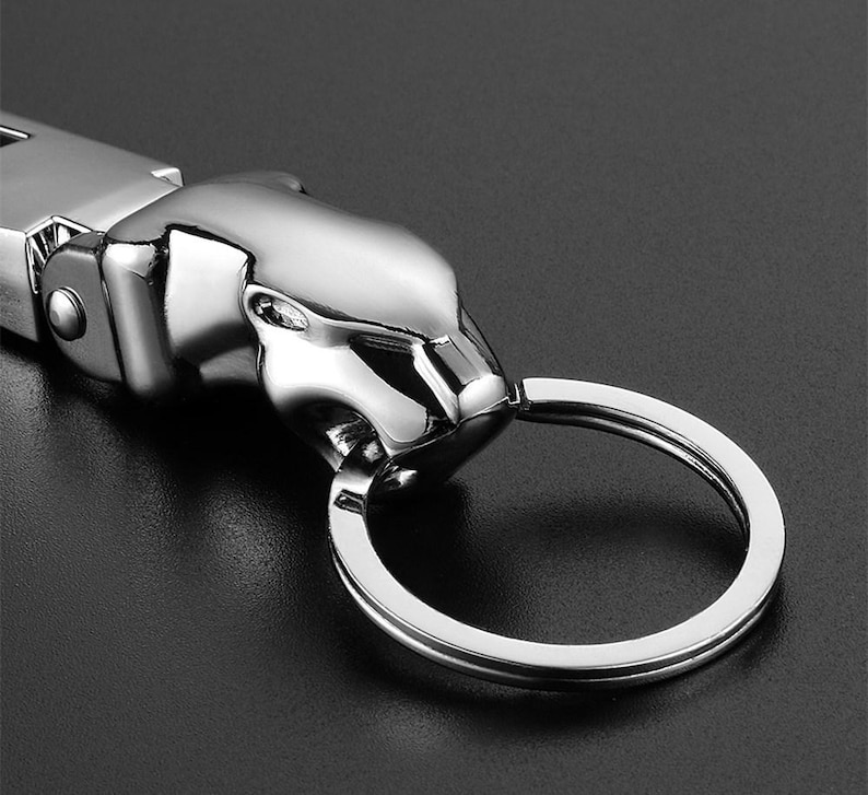 May include: A polished silver-tone metal keychain featuring a stylized jaguar head sculpture. The jaguar head is connected to a circular split ring for keys. The metallic surface has a reflective finish with smooth, contoured features.