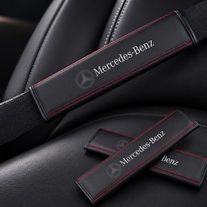 May include: Black seat belt shoulder pads featuring red contrast stitching and the white Mercedes-Benz logo with the three-pointed star emblem. The rectangular pads are designed to wrap around a standard black seat belt strap.