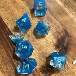 May include: A set of seven polyhedral dice made of translucent blue resin with embedded gear-shaped inclusions. Each die features sharp edges and bright green painted numbers. The set includes a d20, d12, d10, d8, d6, d4, and a percentile die.