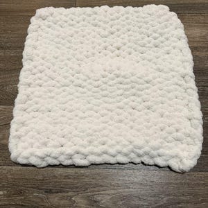 May include: A square, off-white, chunky knit blanket. The blanket is made of a soft, textured yarn, and is lying on a dark wood surface. The blanket's edges are slightly rounded.