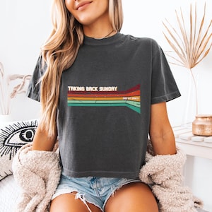 May include: A charcoal gray short-sleeve t-shirt featuring a retro-style graphic with horizontal stripes in red, orange, yellow, and teal. The text "TAKING BACK SUNDAY" is printed in white, with "AND MY SPIRIT" in smaller white text to the right.
