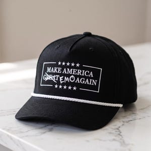 May include: A black baseball cap featuring a white embroidered rectangular border with five stars above and below. The text reads MAKE AMERICA GREAT EMO AGAIN, with the word GREAT crossed out by an X. A white rope trim sits above the brim.