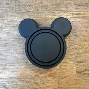 May include: A matte black circular object with two smaller circular protrusions at the top, resembling a stylized head silhouette. The item features a layered, textured surface with a central recessed circle and a raised outer ring, constructed from a solid material.