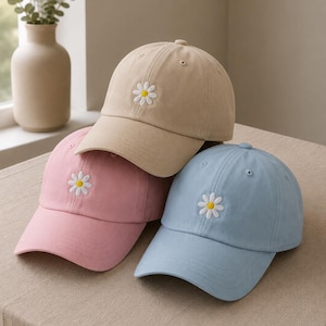 Puede incluir: Tres gorras de béisbol en beige, rosa claro y azul claro, cada una con una pequeña margarita blanca bordada con centro amarillo en el panel frontal. Las gorras tienen una corona estructurada, visera curva y ojales bordados para ventilación.