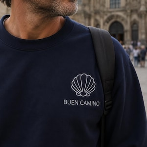 May include: A navy blue crewneck sweatshirt featuring a white embroidered scallop shell icon on the left chest. Below the shell, the text BUEN CAMINO is embroidered in a clean, white sans-serif font. The garment has a ribbed collar and long sleeves.