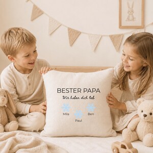 May include: A square off-white throw pillow featuring the black text "BESTER PAPA" and "Wir haben dich lieb" above three light blue snowflake icons. Below each snowflake, the names "Mila", "Paul", and "Ben" are printed in black text.