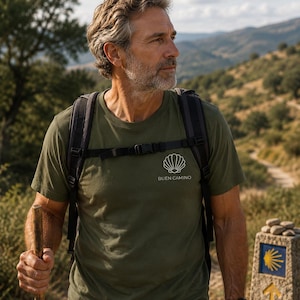 May include: An olive green short-sleeved t-shirt featuring a white line-art graphic of a scallop shell above the text BUEN CAMINO. The shirt is worn with a black backpack, and the design is positioned on the upper left chest area of the garment.