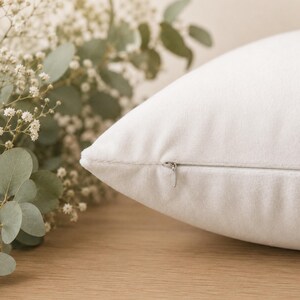 May include: A solid white throw pillow cover featuring a smooth, soft-touch fabric texture and a discreet, concealed zipper closure along the side seam. The pillow is positioned next to a sprig of green eucalyptus and small white flowers.