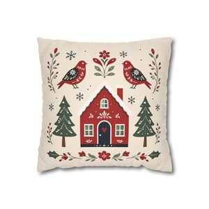 May include: A square throw pillow with a cream-colored background featuring a red house with a dark door and a heart detail, flanked by two green pine trees. The design includes two red birds, floral motifs, and small white snowflakes.