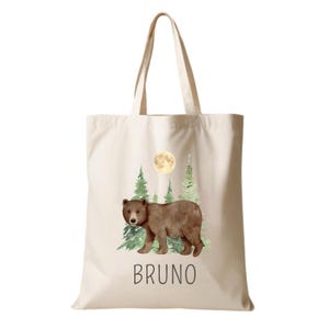 Könnte beinhalten: Eine cremefarbene Stofftasche mit einer Aquarellillustration eines braunen Bären, der vor grünen Kiefern und einem gelben Vollmond steht. Der Name BRUNO ist in schwarzen Großbuchstaben unter der Bärenillustration aufgedruckt.