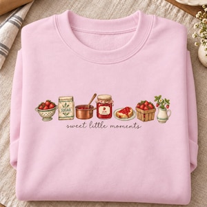 May include: A light pink crewneck sweatshirt featuring a graphic illustration of strawberries, a sugar bag, a copper pot, a jam jar, toast with jam, and a pitcher. The text "sweet little moments" is printed in a cursive font below the illustrations.