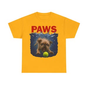 Puede incluir: Una camiseta de manga corta color amarillo mostaza con un estampado gráfico de un perro pequeño de color marrón nadando bajo el agua hacia una pelota de tenis verde neón. La palabra PAWS está impresa en letras rojas sobre la imagen.