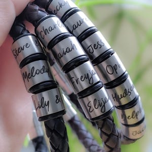 May include: Close-up of multiple leather bracelets with silver beads engraved with names, including Steve, Charlie, Isaiah, Jess, Freya, Opi, Elijah Hudson, Delph, and Jad.