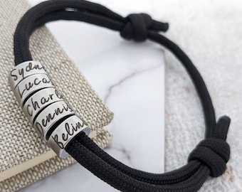 Puede incluir: Pulsera de paracord negro con una pila de cuentas plateadas, cada una grabada con un nombre diferente: Sydney, Luca, Charlie, Dennis y Belin.