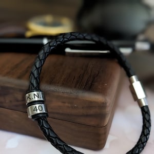 Puede incluir: Pulsera de cuero negro con dos cuentas de metal plateado. Las cuentas están grabadas con las letras "K.N." y el número "40".