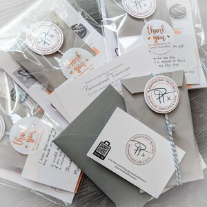K&ouml;nnte beinhalten: Mehrere verpackte Artikel, darunter kleine graue Umschl&auml;ge, Karten und transparente Plastikt&uuml;ten, sind auf einer hellen Oberfl&auml;che angeordnet. Jedes Paket ist mit einem runden Aufkleber mit der Aufschrift "Danke" und dem Logo des Shops versehen.