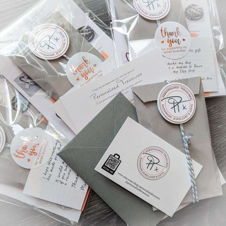 K&ouml;nnte beinhalten: Mehrere verpackte Artikel, darunter kleine graue Umschl&auml;ge, Karten und transparente Plastikt&uuml;ten, sind auf einer hellen Oberfl&auml;che angeordnet. Jedes Paket ist mit einem runden Aufkleber mit der Aufschrift "Danke" und dem Logo des Shops versehen.