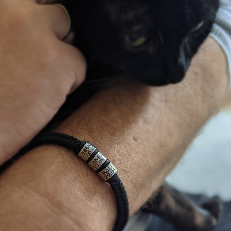 K&ouml;nnte beinhalten: Schwarzes geflochtenes Armband mit drei silbernen Perlen, die mit Namen und Daten graviert sind. Das Armband wird am Handgelenk einer Person getragen, mit einer schwarzen Katze im Hintergrund.