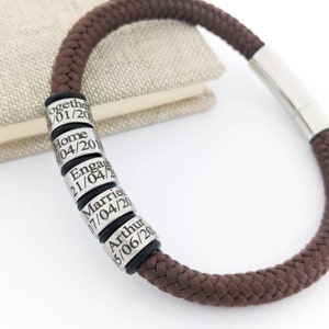 K&ouml;nnte beinhalten: Braunes geflochtenes Seilarmband mit silberfarbenen zylindrischen Perlen. Jede Perle hat gravierten Text und Daten. Das Armband hat einen Magnetverschluss. Im Hintergrund ein Buch in neutraler Farbe.