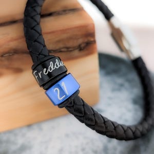 May include: Black leather braided bracelet with a silver clasp. The bracelet has two engraved metal beads, one with the name "Freddie" and the other with the number "21".